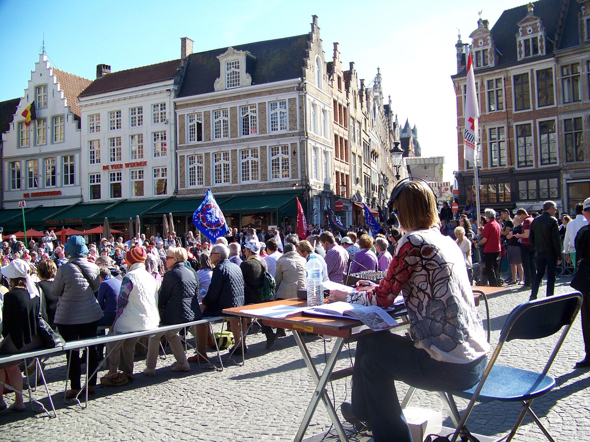 Susanne zit aan een tafel op een klein podium achter de rijen met bankjes voor het publiek. Op de achtergrond trekt de processie voorbij. Een groep vendelzwaaiers houdt metershoge gekleurde vlaggen op.