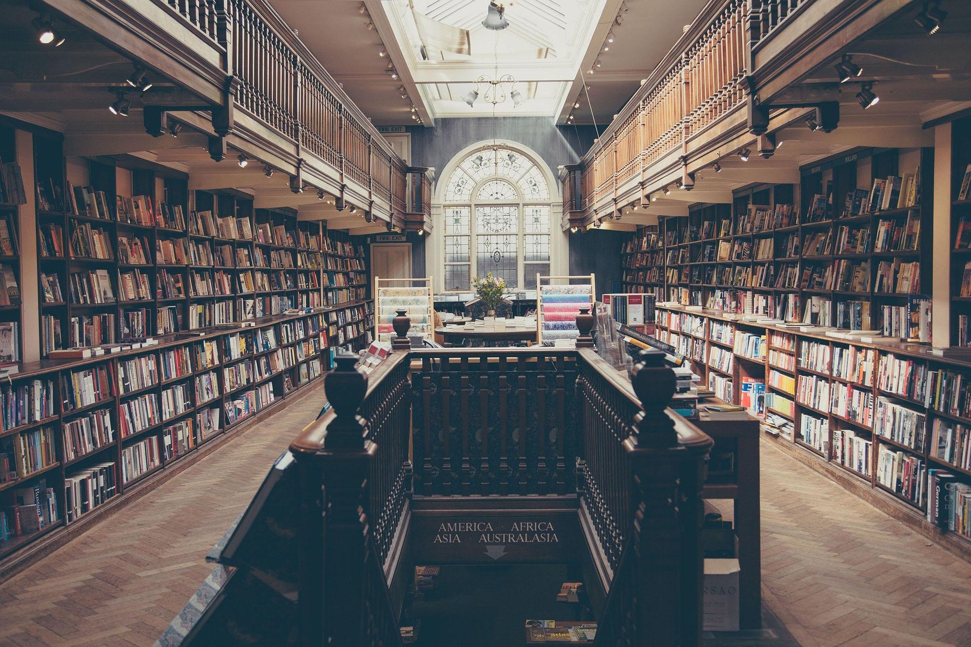 Een bibliotheek met donkere, houten boekenkasten en een houten vloer in visgraatmotief. Op de achtergrond een groot boograam.